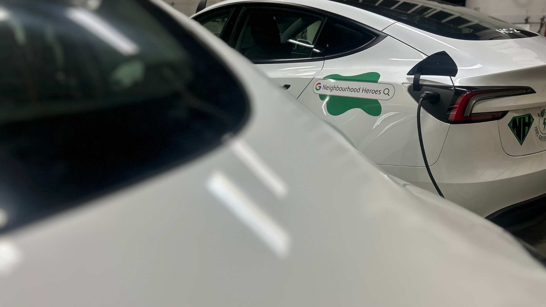 White Tesla Model 3 charging inside a garage with Neighbourhood Heroes branding on the rear door and another vehicle blurred in the foreground.
